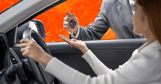 A man in a gray suit hands a key to a woman in a white sweater seated in the driver's seat of a car.