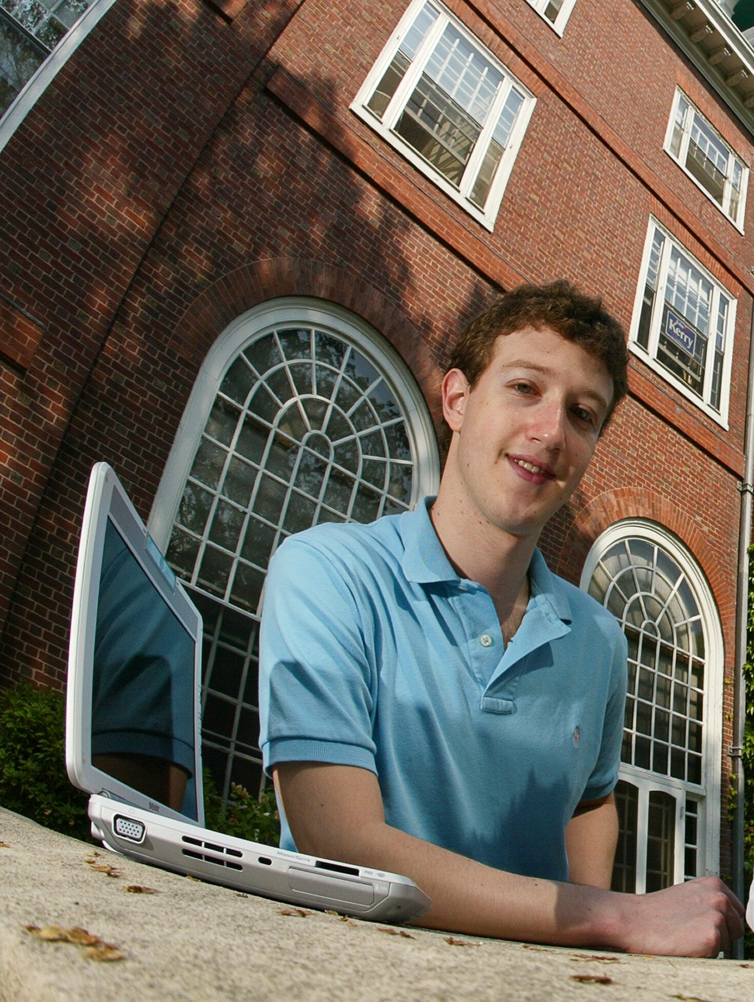 A young Mark Zuckerberg smiling outdoors while leaning over an open laptop in front of a brick building.