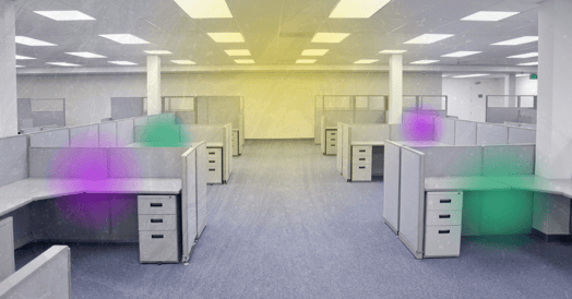 An interior view of an empty office space filled with rows of grey cubicles. Overhead fluorescent lights illuminate the scene.