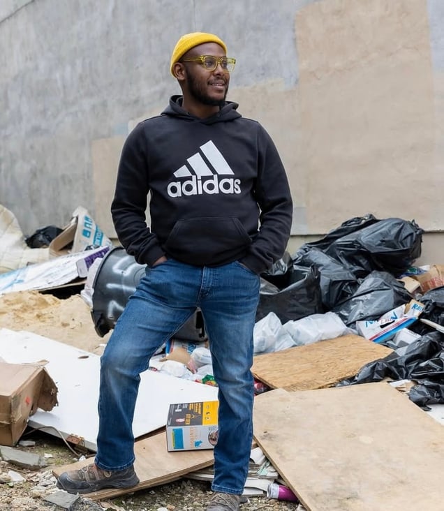 Terrill Haigler, known as "Ya Fav Trashman," standing in a yellow beanie and Adidas hoodie in front of a pile of trash bags.