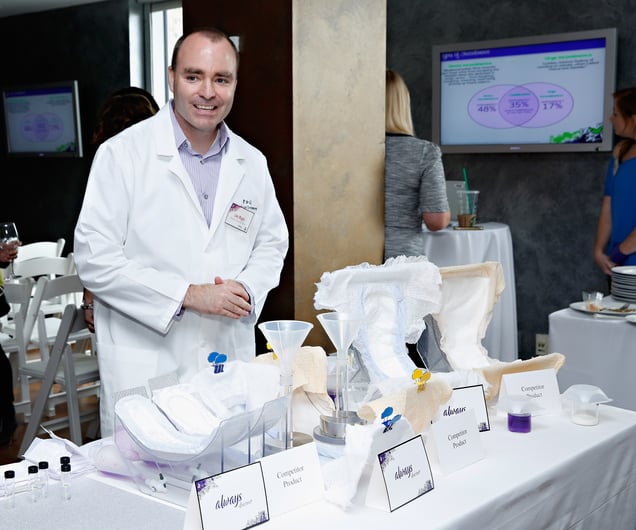 A photograph of a man in a white lab coat standing behind a table displaying various "Always" brand feminine hygiene products at a promotional event.