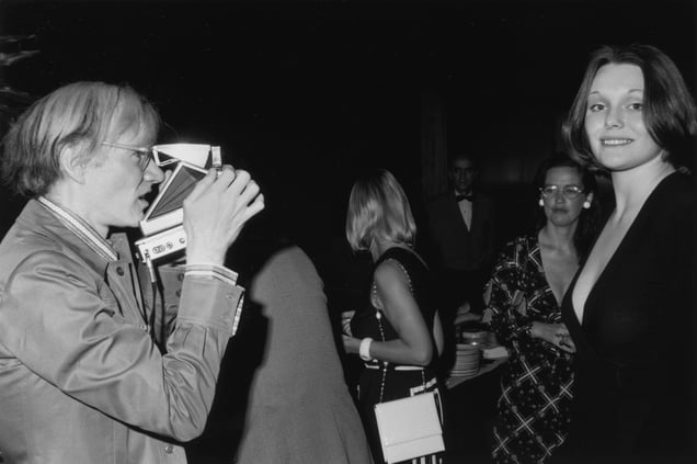 A black-and-white photo from 1973 showing artist Andy Warhol using a Polaroid camera to photograph model Tessa Dahl at a party.