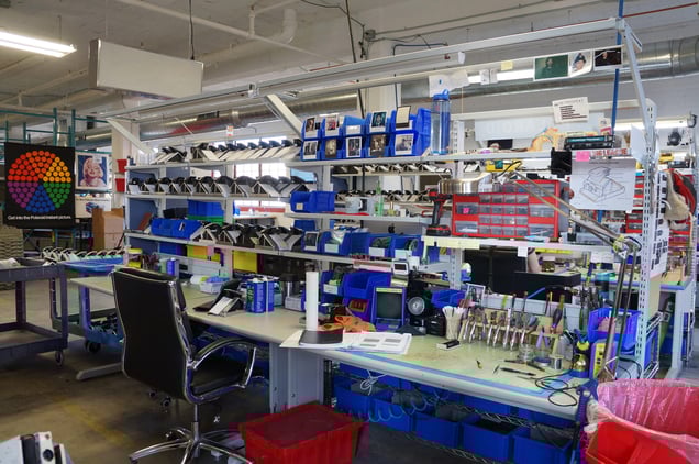 An interior view of the Milwaukee workshop featuring long workbenches lined with blue bins and various refurbished electronics.