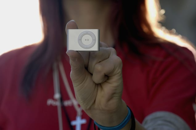 A person's hand holding a tiny, silver second-generation iPod Shuffle between their thumb and forefinger.