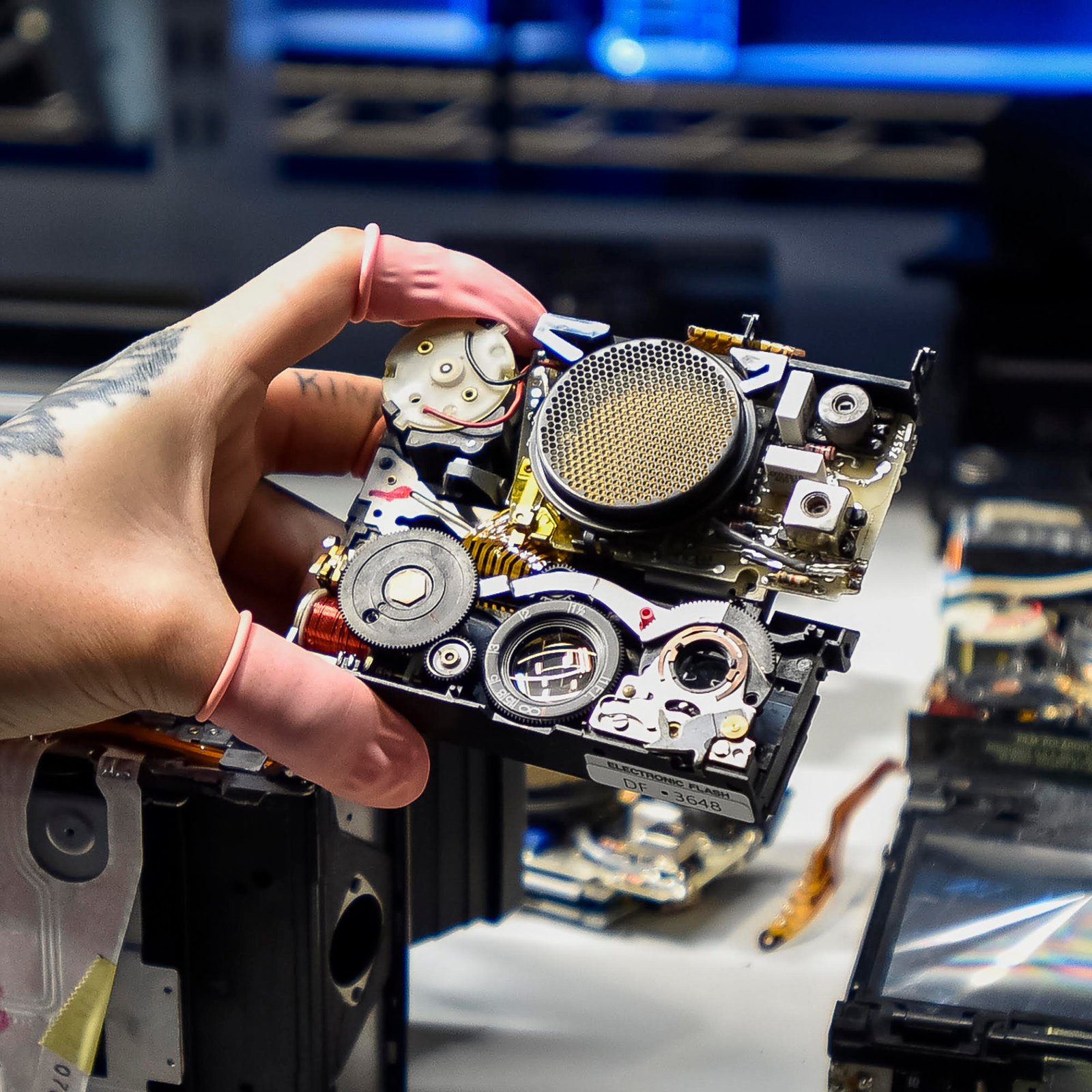 A close-up shot of a technician's hand holding the intricate internal components and circuitry of a disassembled Polaroid camera.