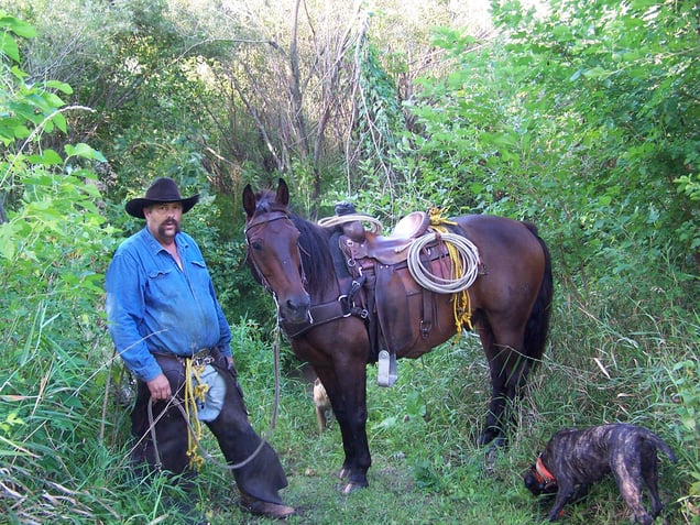 Chet Peugh and Horse: A man with a mustache and a cowboy hat (Chet Peugh) stands in a wooded area next to a saddled brown horse and a brindle dog.