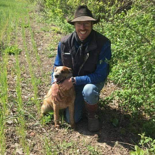 Wesley Bush and Dog: A smiling man in a cowboy hat and vest (Wesley Bush) kneels on a dirt path next to a medium-sized tan dog.