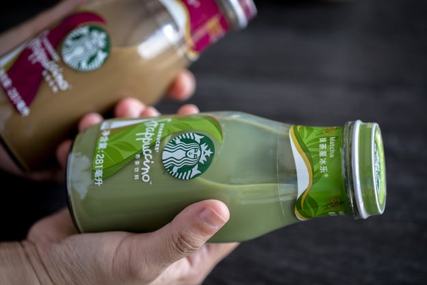 A person holds a glass bottle of Starbucks Matcha Frappuccino, with the green label and Starbucks logo clearly visible. Another similar bottle, slightly out of focus, is in the background.