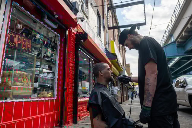 Man receiving a haircut from a barber outside a shop on a busy Kensington Avenue sidewalk.