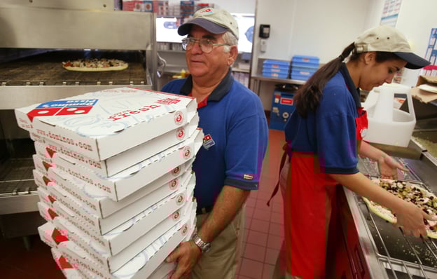 Pizza shop scene: one person in blue shirt stacking white boxes with red text, another in red apron preparing pizza; red tiled floor and pizza oven in background.