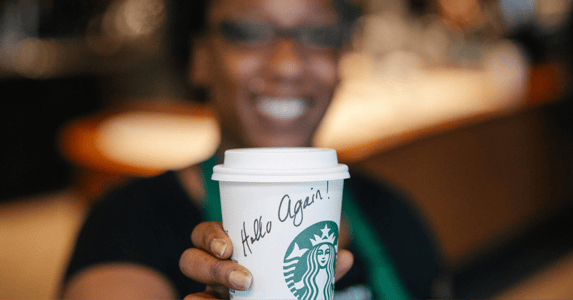A smiling Starbucks barista, wearing a green apron, holds out a white paper cup with the Starbucks logo and the handwritten message 