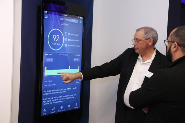 Two men in suits look at and point to a large, vertical digital display showing a sleep tracking app interface, which has a sleep score of 92.