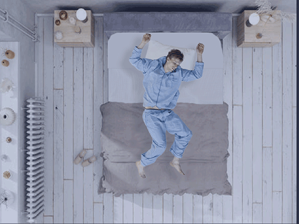 An overhead shot of a man wearing blue pajamas lying on a bed with his arms raised, as if struggling with sleep.