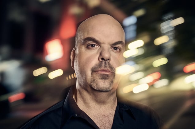 A close-up portrait of Johnathan Walton, a bald man with a goatee, standing outside at night with city lights blurred in the background.