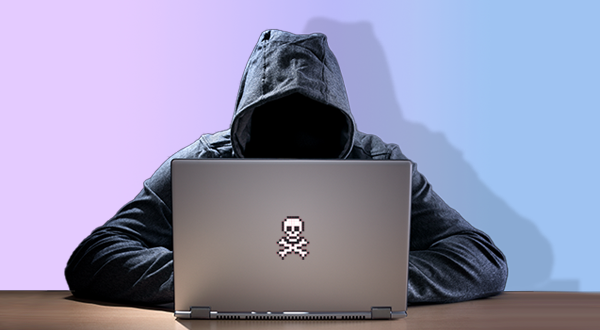 A person in a black hooded sweatshirt sits in front of a laptop with a pixelated skull and crossbones logo on the lid, suggesting a hacker or cyber criminal.
