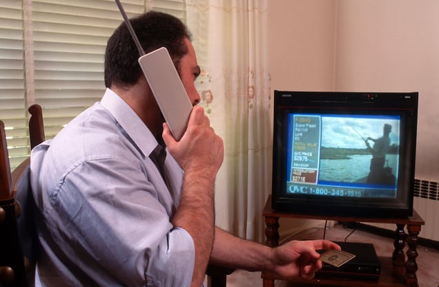 A man on an early cell phone holds a credit card, while a television in the background displays an image of a person fishing with a shopping channel overlay.