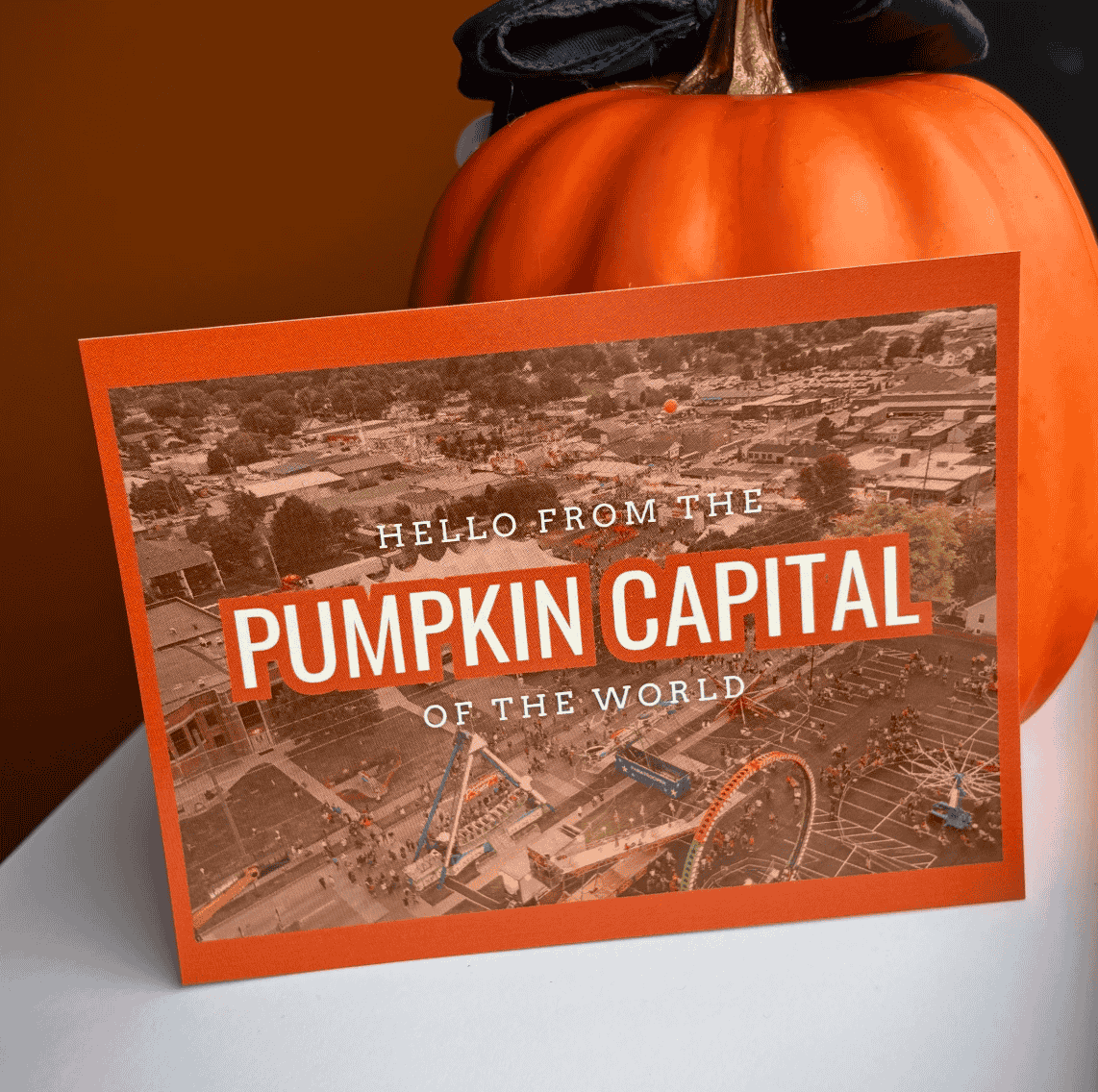 A postcard-style image that reads "HELLO FROM THE PUMPKIN CAPITAL OF THE WORLD" with an aerial photo of a town and a pumpkin festival on it, propped up next to a pumpkin.