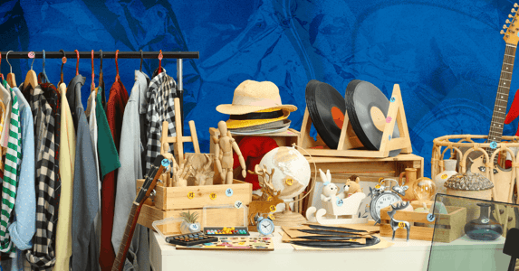 A rack of clothes next to a table piled with items including vinyl records, a globe, a guitar, and various knick knacks. 
