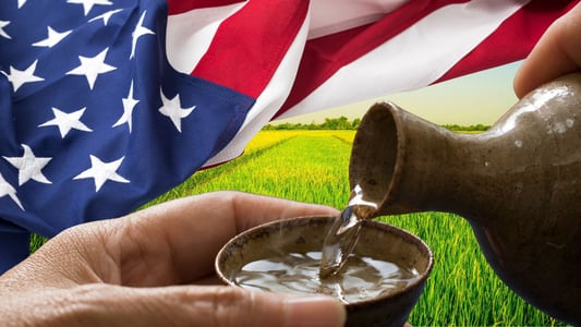 Two hands for sake into a cup in front of a green field and an American flag. 