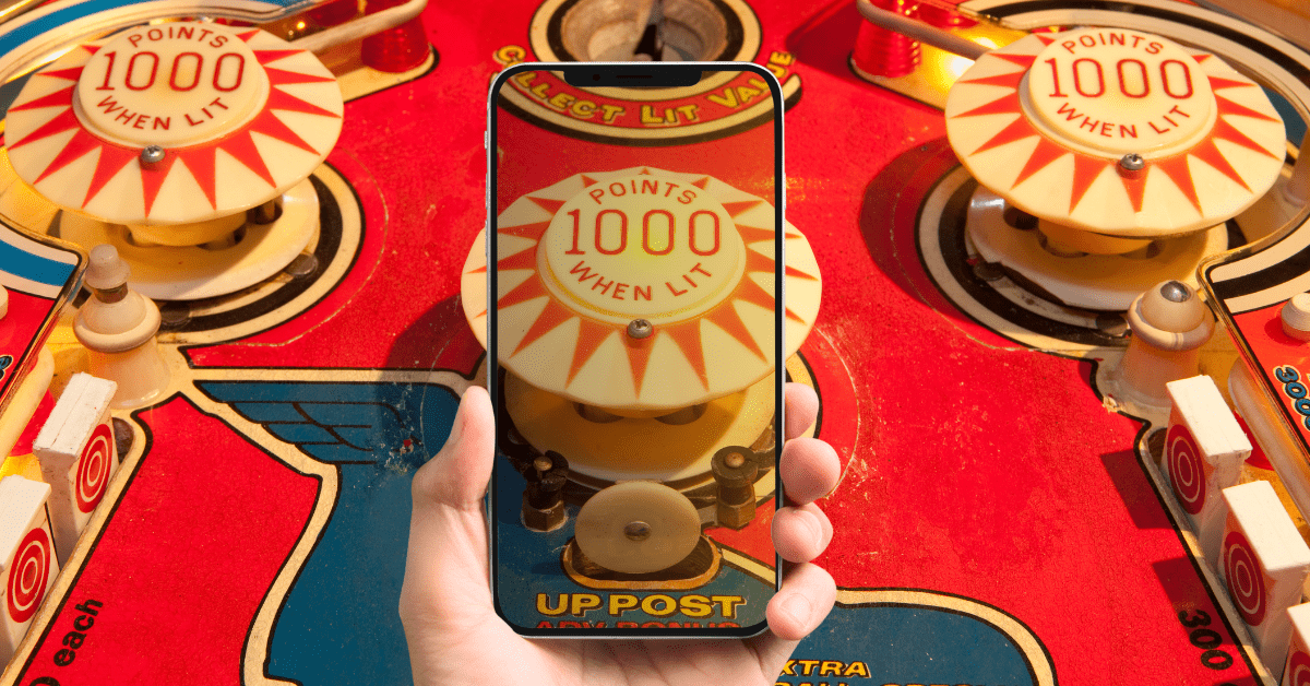 A pinball machine with a hand holding a smartphone in the foreground displaying the same machine on its screen.