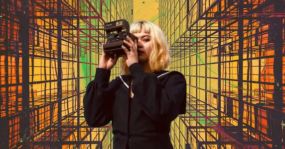 A woman with short blonde hair wearing a black dress takes a photo with a black camera on a yellow, green, and orange background with a geometric pattern on it.