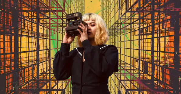A woman with short blonde hair wearing a black dress takes a photo with a black camera on a yellow, green, and orange background with a geometric pattern on it.