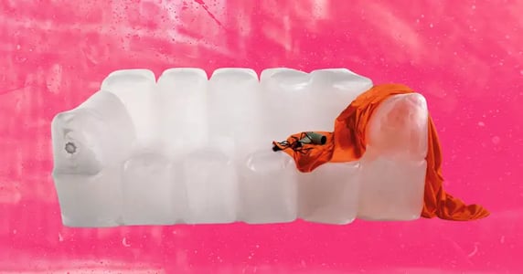 A clear, inflatable couch on a pink background. A piece of orange fabric is draped over one arm, and a green hair dryer sits on a cushion.
