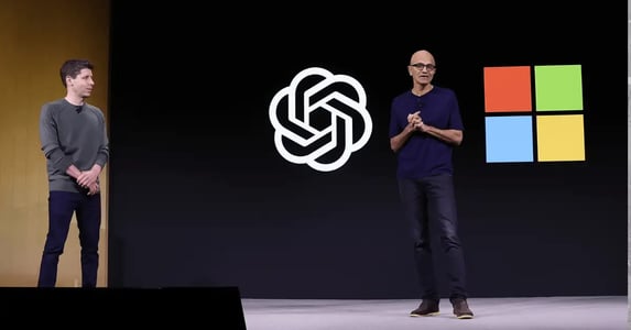 Microsoft CEO Satya Nadella speaks on stage with OpenAI and Microsoft logos displayed on a screen behind him. OpenAI CEO Sam Altman stands to the left, looking on.