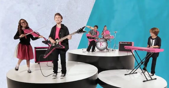 Five kids wearing pink and black outfits perform on raised platforms as a band.