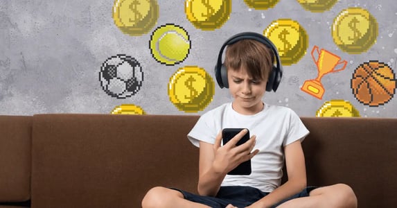 A little boy sitting cross-legged on a brown couch with black headphones staring at a smartphone while pixelated coins and sports-related symbols fall in the background.