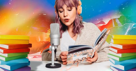 A woman with pink hair reading a book while wearing pink headphones and speaking into a silver microphone with stacks of rainbow books on either side of her desk and a rainbow background.