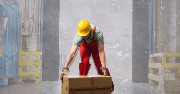 A man in red overalls and a yellow construction hat bends down to lift a brown cardboard box.
