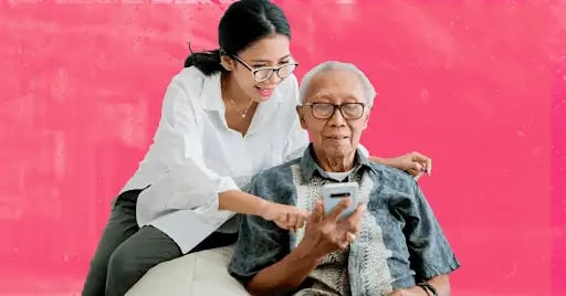 An Asian woman in a white button-down and glasses helping an elderly Asian man with white hair and glasses look at someone on a smartphone.