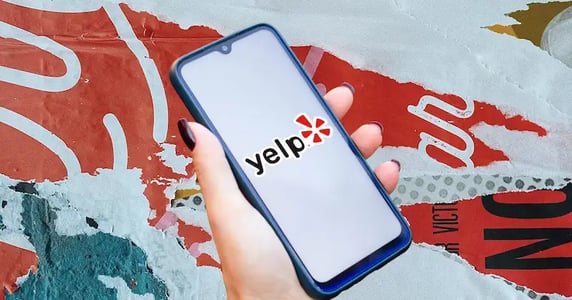 A hand holding a phone with the Yelp logo on the screen over a background of torn paper.