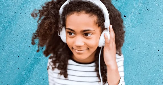A young girl with curly hair wearing over-ear headphones and smiling on a blue background.