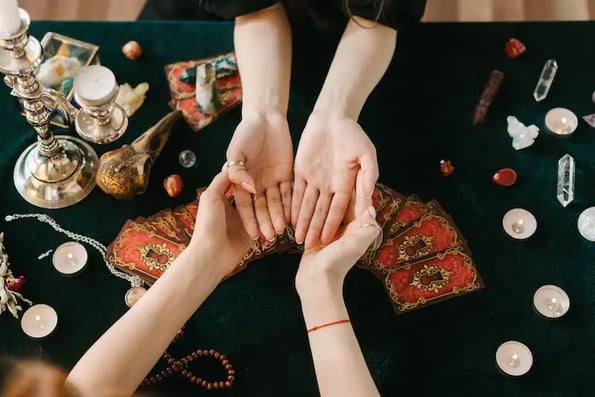 A woman examines another woman’s palms over a table covered in candles, crystals, and tarot cards.