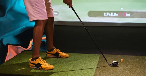 A man from the waist down in pink shorts and yellow sneakers teeing up at an indoor golf hole with a screen in the background.