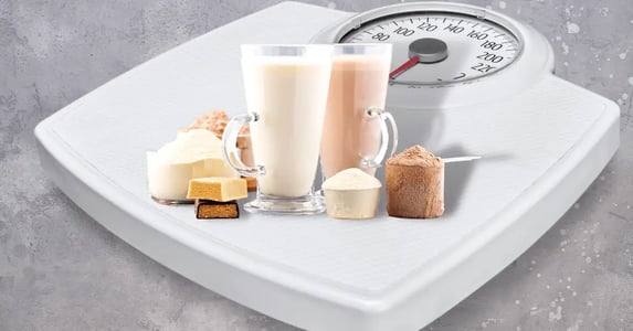 A weight scale with protein shakes, powder, and bars stacked on top of it on a gray background.