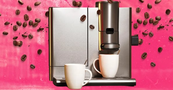 A silver espresso machine with two white mugs on a pink background with coffee beans falling from the top.