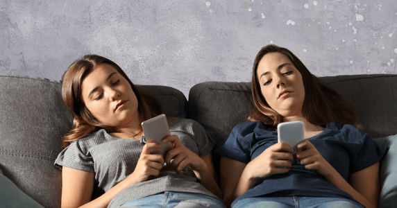 Two young women laying on a couch looking at their phones.