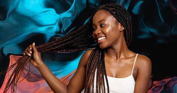 A Black woman in a white tank top smiles while holding a section of her braided hair.