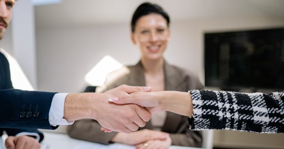 A man and a woman shake hands. In the background, another woman is smiling. 
