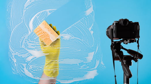 A hand wearing a yellow dish glove scrubs with a soapy sponge while a video camera records.