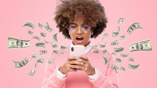 A young Black woman in a pink sweater and glasses looks at her phone in disbelief as money flies out.