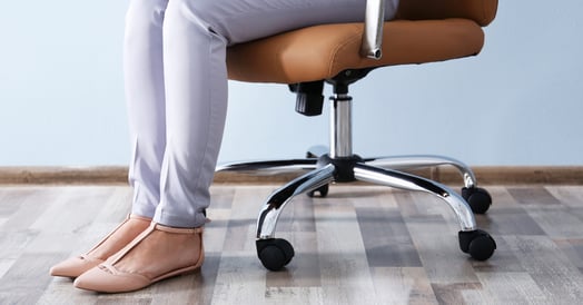 A woman's legs are visible as she sits in a desk chair with wheels. She wears light-colored slacks and tan shoes.