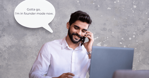 A man sitting in front of two laptops is talking on a cellphone, saying, “Gotta go, I’m in founder mode now.”