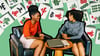 Two women chat at a small table with a laptop and on it. Behind them are several Mahjong tiles.