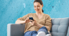 A woman sits on the couch and smiles as she looks at her phone.