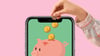 A child’s hand putting a coin into a piggy bank displayed on a smartphone screen. 