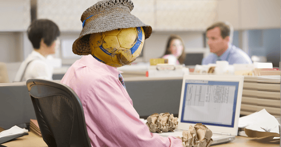A seemingly human body sitting in front of a computer in an office, but the head is a soccer ball wearing a hat and the hands are wads of paper.